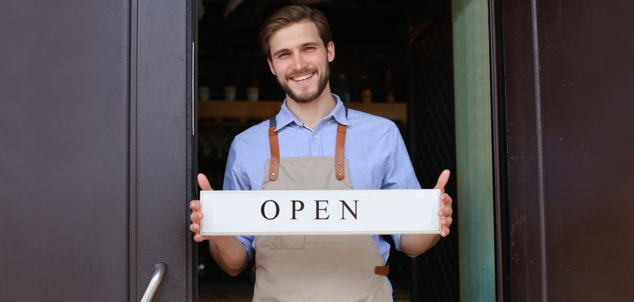 Smiling male owner of coffee shop is standing outdoors. He is holding placard and inviting everybody to come in. Open.